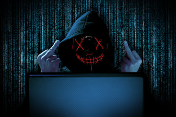 hacker with middle fingers gesture provocation red glowing mask behind notebook laptop in front of...