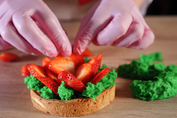 Obraz na płótnie Canvas Pastry chef is putting strawberry slices with cream on biscuit making a mini cake. Hands close-up. Baking and industrial food production