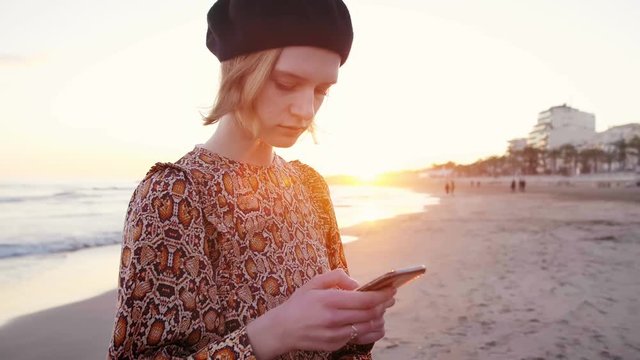 Blond girl surprised to get message on the phone standing on the beach in sunset light. Middle side shot of gorgeous girl in beret and stylish dress near sea checking mail,