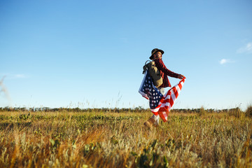 4th of July. Fourth of July. American with the national flag. American Flag. Independence Day. Patriotic holiday. The man is wearing a hat, a backpack, a shirt and jeans. Beautiful sunset light. 