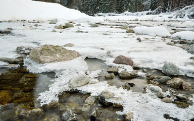 Shallow forest river covered with crystal snow and ice layer in winter, white patches over stones, long exposure photo