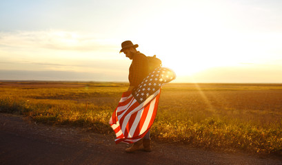 4th of July. Fourth of July. American with the national flag. American Flag. Independence Day. Patriotic holiday. The man is wearing a hat, a backpack, a shirt and jeans. Beautiful sunset light. 