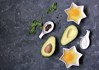 Ingredients for cooking baked avocado with egg on a dark concrete background: ripe avocado, cut in half, eggs, salt, pepper, parsley. Top view, copy space.