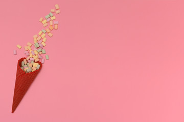 Red waffle ice cream cone. The scattered multi-colored marshmallows isolated on a pink background. Congratulations, celebration concept. Top view. Copy space.
