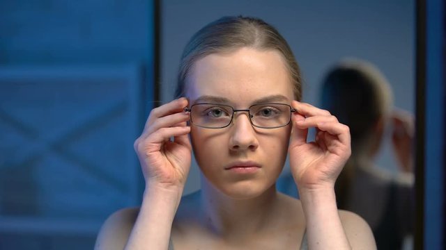 Upset female teenager trying on eyeglasses, unsatisfied with mirror reflection