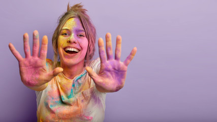 Indoor shot of glad positive Caucasian woman shows colorful palms, stretches hands, splashes powder, smiles positively, isolated over purple background with empty space. Holi festival concept