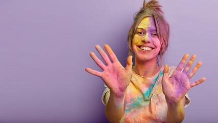 Happy Holi day concept. Funny satisfied girl with colorful powder on face, clothes and palms, being in high spirit, celebrates traditional spring festival, poses against purple wall with free space