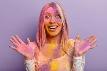 Headshot of glad female with toothy smile, happy reaction, spreads hands, dirty with multicolored...