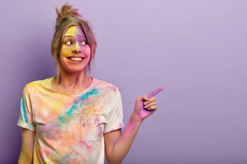 Merry delighted female plays with holi colors, has fun at festival, points away with fore finger,...