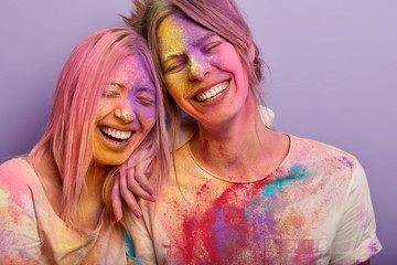 Sincere emotions and feelings concept. Funny two female friends lean heads to each other, laugh gladfully, have broad smiles, colored dirty faces, splattered clothes, participate in Holi festival