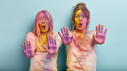 Overwhelmed two girls have surprised facial expressions, show colorful palms at camera, open mouth from shock, asks not shower them with powder, painted with colors, isolated over blue background