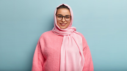 Half length shot of beautiful dark eyed smiling female in pink jumper and hijab, looks through...