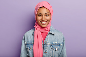 Studio shot of glad Eastern female has Islamic religion, covered head with pink veil, smiles gently, shows white teeth, isolated against violet wall expresses positive feelings and emotions. Ethnicity - obrazy, fototapety, plakaty