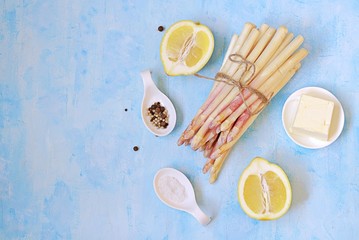 Ingredients for cooking asparagus on a light blue concrete background: fresh white asparagus, lemon, butter, salt, pepper. Top view, copy space.