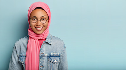 Glad satisfied delighted woman smiles broadly at camera, being in high spirit, looks through optical glasses, has Islamic views, stands against blue studio wall with empty space for your promotion - obrazy, fototapety, plakaty