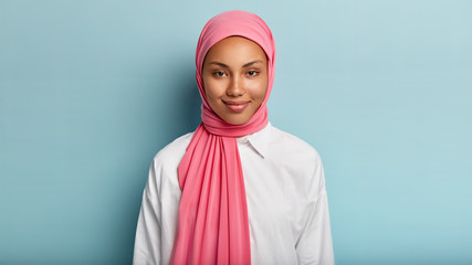 Headshot of lovely satisfied religious Muslim woman with gentle smile, dark healthy skin, wears pink scarf on head, white shirt, isolated over blue background, has no make up, natural beauty