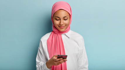 Satisfied ethnic female focused in smartphone device, checks newsfeed online, enjoys using modern gadget, wears pink veil, elegant white shirt, has gentle smile on face, isolated over blue wall