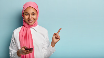 Photo of glad Arabic woman with gentle smile on face, points with fore finger on blank space, pink veil on head, holds mobile phone for texting messages, isolated against blue wall, browses internet