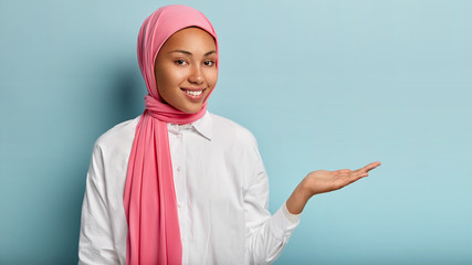 Satisfied beautiful woman teaches Muslim religion, raises palm over blank space as if holding something, wears tradition hijab, white clothes, isolated over blue studio wall. Advertisement concept