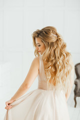 Beautiful blonde woman with greek hairstyle in beige powdery atlas wedding dress posing in studio room.