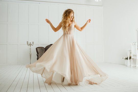 Beautiful Blonde Woman With Greek Hairstyle In Beige Powdery Atlas Wedding Dress Posing In Studio Room.