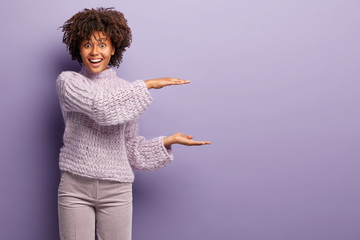 Studio shot of of glad Afro American woman does size gesture, demonstrates something large, has...