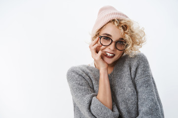 Waist-up shot of modern stylish good-looking european female with blond hairstyle in glasses warm winter beanie and sweater biting finger flirty and cheeky making romantic gazes over white wall