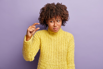 Puzzled beautiful Afro American woman shows small amount, little gesture, demonstrates tiny size, dissatisfied with something no very big, dressed in casual yellow sweater, poses indoor. Hand gesture