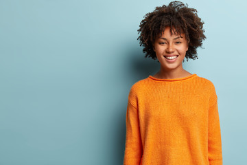 Photo of lovely positive Afro American woman with curly hair, wears orange jumper, being in high...