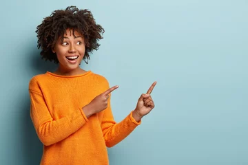 Muurstickers Horizontal shot pleased dark skinned female with Afro haircut, points away with both fore fingers, shows blank space for your promotion, isolated over blue background. People, advertisement concept © Wayhome Studio