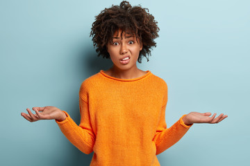 Horizontal shot of attractive woman with puzzled expression, spreads hands with confused look, frowns face, wears orange clothes, feels hesitant, indignant isolated over blue background. Doubt concept