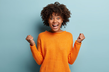 Joyful dark skinned model with crisp hair, clenches fists, feels overjoyed after winning game,...