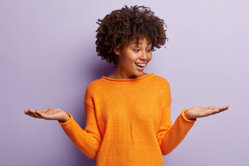 Glad optimistic Afro American woman raises both palms, pretends holding two items, being in good...