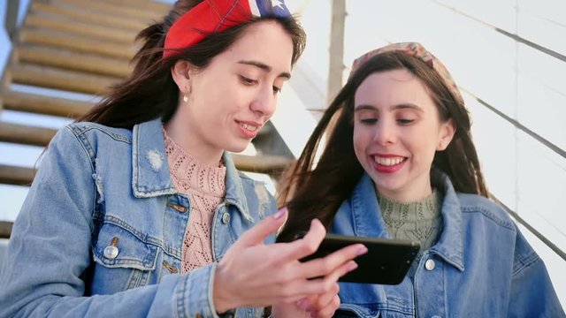 Two nice young Hispanic sisters exploring new app on smart phone device, stays connected and up to date, texting messages to family and friends, while sit on stairs in urban area, dressed in jeans
