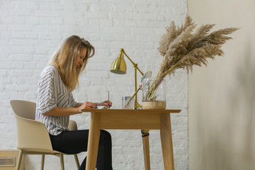 side view of young woman with blond hair, striped blouse, handwriting some notes at the wooden...