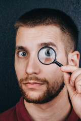 Fototapeta na wymiar a young man with a beard looks through a magnifying glass. Portrait of a guy with a big eye on a black background. investigation, survey