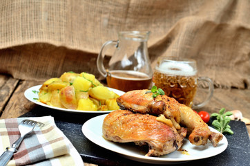 Golden grilled chicken legs on a plate, roasted potatoes and jug of cold beer, rosemary, tomatoes, oregano, salt, pepper and garlic on a wooden table