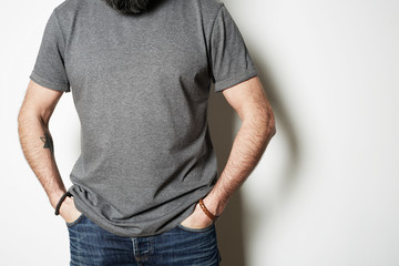 Obraz premium Attractive brutal tattooed bearded guy poses in blue jeans and blank gray t-shirt premium summer cotton, on white background mockup.