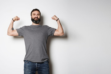 Positive brutal tattooed bearded guy poses in blue jeans and blank gray t-shirt premium summer cotton, on white background mockup.