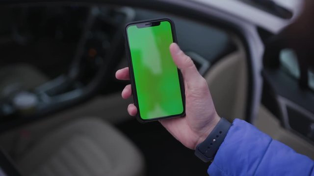 Hand of man wearing in blue jacket holding smartphone with green screen. Guy sitting in automobile. Vehicle. Device, digital technology, technology, traffic jam, transport, transportation, travel