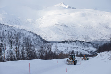 Fototapeta na wymiar Snow plow truck clearing icy road after winter snowstorm blizzard for vehicle access Snow blower clears snow-covered streets producing a plume of snow. Winter landscape in Norway or Sweden Scandinavia