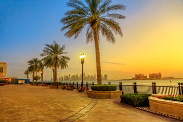 Benches and palm trees along marina walkway in Porto Arabia at the Pearl-Qatar, Doha, with skyscrapers of West Bay skyline illuminated at blue hour.Scenic sunset landscape of Persian Gulf, Middle East © bennymarty