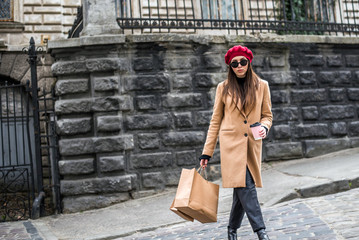 Attractive cute girl in with the shopping bags and warm drink in her hand. Young beautiful woman with long brown hair in sunglasses on a walk on a cloudy day. Fashionable sexy girl. 