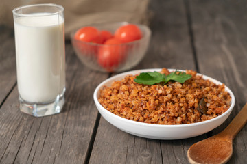 Buckwheat porridge with a sprig of parsley in a bowl with a glass of milk and tomatoes on a dark rough wooden table. traditional Russian dish. Rustic style. Copy space.