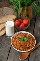 Buckwheat porridge with a sprig of parsley in a bowl, with a glass of milk and tomatoes on a dark rough wooden table. traditional Russian dish. Rustic style. Copy space.