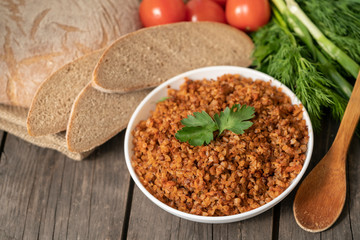 Buckwheat porridge with a sprig of parsley in a bowl, with a glass of milk and tomatoes on a dark rough wooden table. traditional Russian dish. Rustic style. Copy space. dill, onion, greens, top view