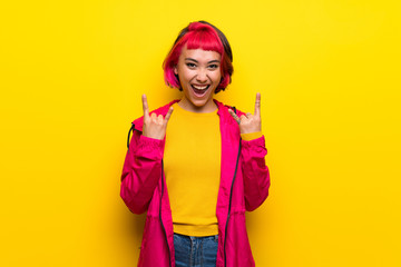Young woman with pink hair over yellow wall making rock gesture