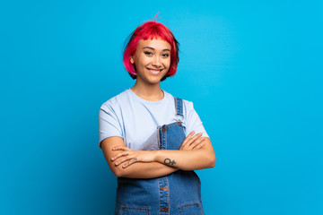 Young woman with pink hair over blue wall keeping the arms crossed in frontal position