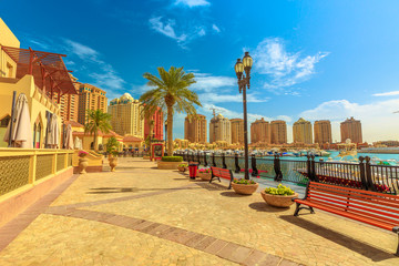 Benches and palm trees along marina walkway promenade in Porto Arabia at the Pearl-Qatar, Doha, with residential buildings towers on background. Persian Gulf in Middle East. Sunny day, blue sky.