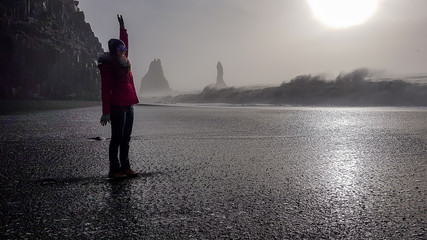A young woman wearing a pink jacket, standing at a wet beach at Reynisfjara Black Sand Beach, Iceland, with one of her arm up in the air. Having fun with the waves. Joy and happiness.